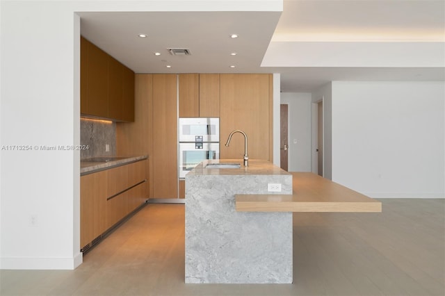 kitchen with white double oven, tasteful backsplash, sink, and an island with sink