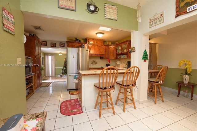 kitchen featuring kitchen peninsula, stainless steel fridge, a breakfast bar area, decorative backsplash, and light tile patterned floors