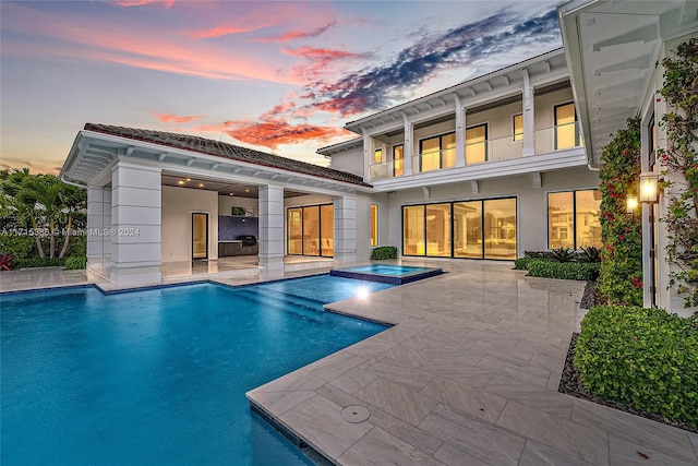 back house at dusk featuring a swimming pool with hot tub, a balcony, and a patio