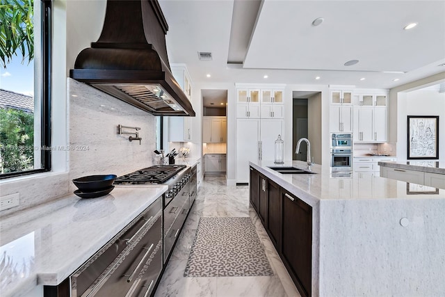 kitchen with custom exhaust hood, a large island with sink, sink, white cabinetry, and stainless steel appliances