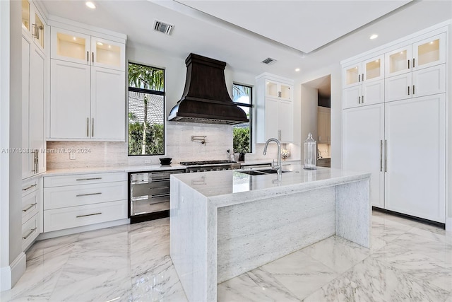 kitchen with custom exhaust hood, a kitchen island with sink, sink, light stone countertops, and white cabinetry