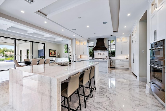 kitchen with white cabinetry, a raised ceiling, light stone counters, premium range hood, and a spacious island