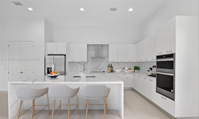 kitchen featuring tasteful backsplash, white cabinets, a kitchen breakfast bar, stainless steel appliances, and wall chimney range hood