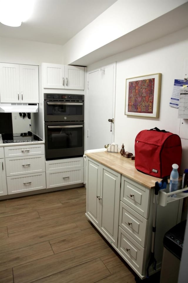 kitchen with white cabinets, black electric stovetop, multiple ovens, under cabinet range hood, and wooden counters