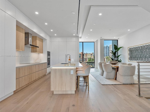 kitchen featuring white cabinets, wall chimney range hood, a kitchen island with sink, and light hardwood / wood-style flooring