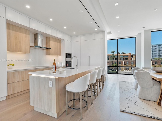 kitchen featuring light wood-type flooring, wall chimney exhaust hood, a kitchen island with sink, a wall of windows, and white cabinets