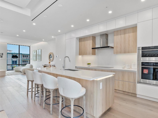 kitchen with sink, wall chimney range hood, double oven, a large island with sink, and white cabinets