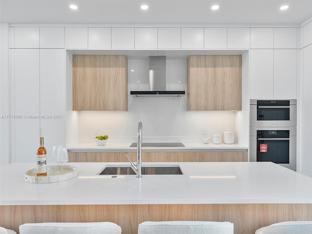 kitchen featuring white cabinetry, sink, stainless steel double oven, wall chimney range hood, and a kitchen breakfast bar