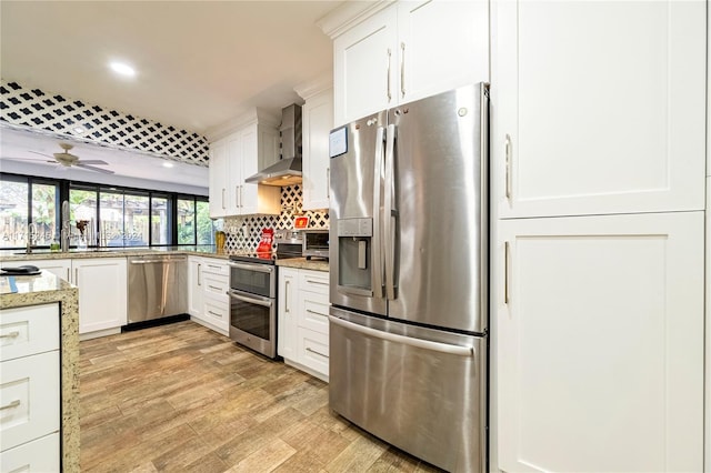 kitchen with wall chimney exhaust hood, ceiling fan, decorative backsplash, appliances with stainless steel finishes, and white cabinetry