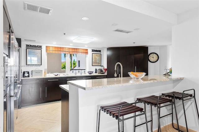 kitchen with a kitchen breakfast bar, kitchen peninsula, light stone countertops, and dark brown cabinetry