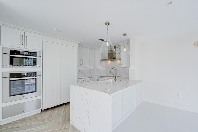 kitchen with white cabinetry, stainless steel double oven, wall chimney range hood, light stone counters, and decorative light fixtures