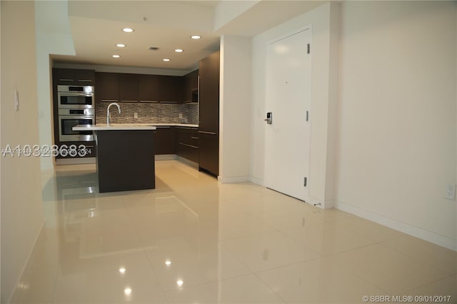 kitchen with backsplash, double oven, an island with sink, a breakfast bar area, and light tile patterned floors