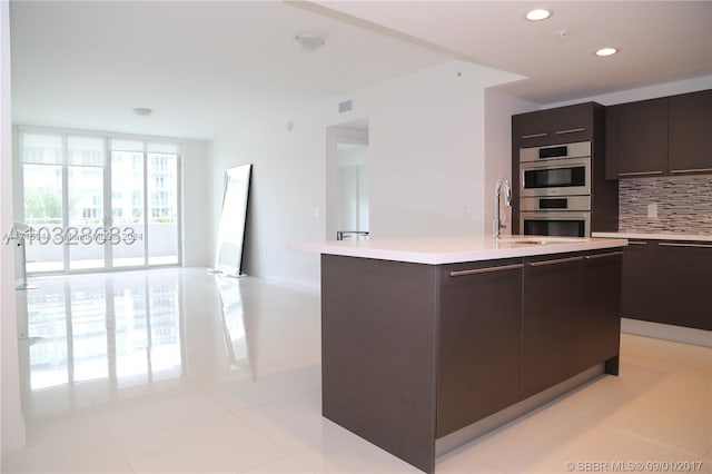 kitchen featuring floor to ceiling windows, backsplash, a center island with sink, light tile patterned floors, and stainless steel double oven