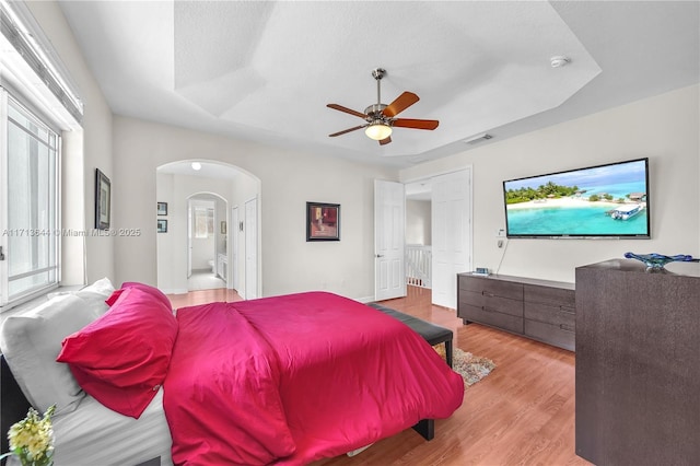 bedroom with a tray ceiling, visible vents, arched walkways, light wood-style floors, and ceiling fan