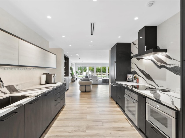 kitchen with stainless steel oven, sink, light stone countertops, range hood, and light hardwood / wood-style floors