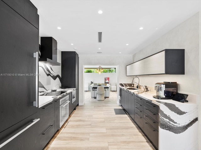 kitchen featuring stainless steel oven, sink, light stone counters, and wall chimney range hood