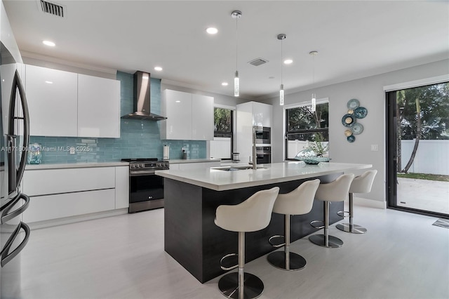 kitchen featuring white cabinetry, wall chimney exhaust hood, an island with sink, pendant lighting, and appliances with stainless steel finishes