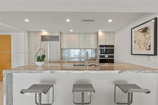 kitchen with light stone countertops, sink, a kitchen breakfast bar, black double oven, and paneled built in refrigerator