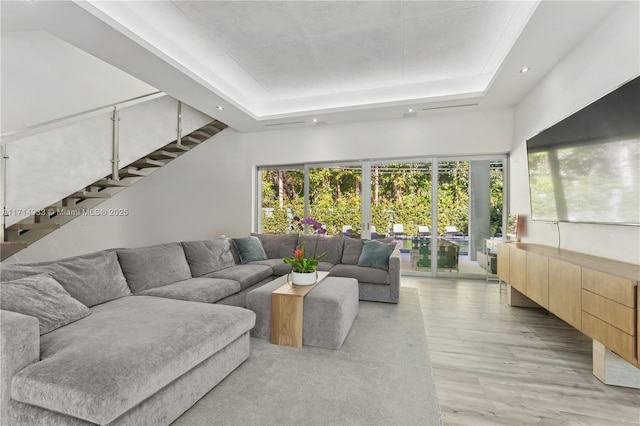 living area with plenty of natural light, stairs, light wood-style flooring, and a raised ceiling