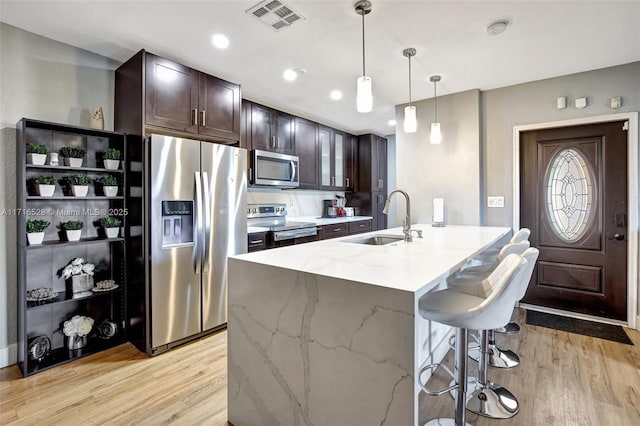 kitchen with light stone counters, stainless steel appliances, a kitchen island with sink, sink, and decorative light fixtures