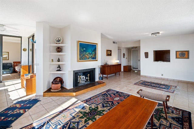 tiled living room with a multi sided fireplace, built in shelves, ceiling fan, and a textured ceiling