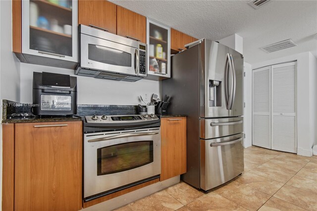 kitchen featuring a textured ceiling, light tile patterned floors, appliances with stainless steel finishes, and dark stone countertops