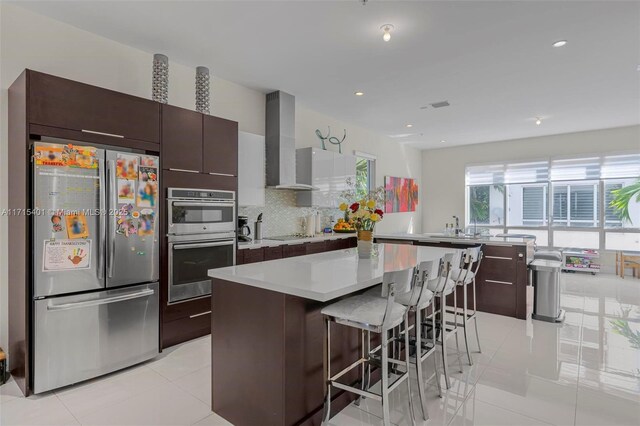 kitchen featuring wall chimney exhaust hood, stainless steel appliances, backsplash, a breakfast bar, and a kitchen island