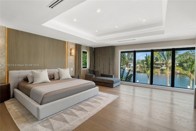 bedroom featuring light hardwood / wood-style floors, a water view, multiple windows, and a tray ceiling