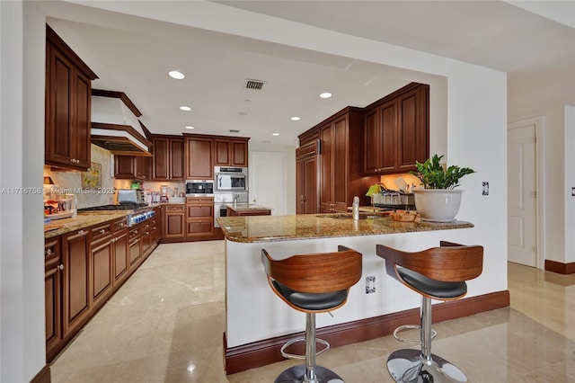 kitchen with a kitchen bar, kitchen peninsula, multiple ovens, stone countertops, and stainless steel gas stovetop