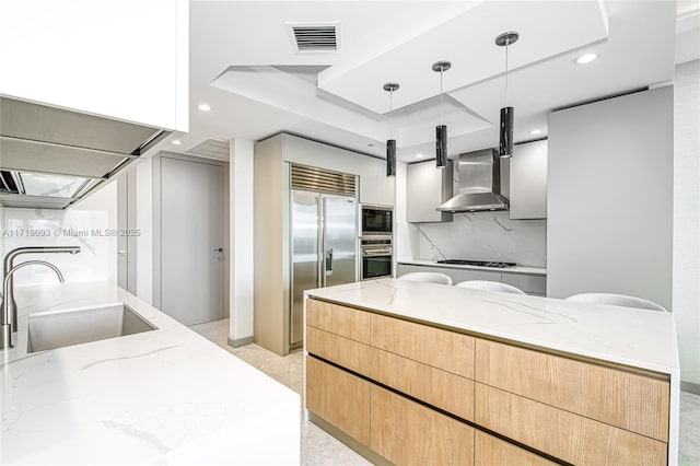 kitchen featuring sink, built in appliances, wall chimney exhaust hood, decorative light fixtures, and light stone counters