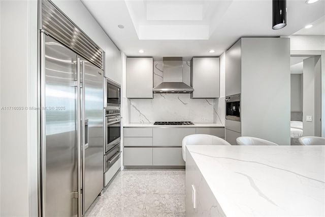 kitchen featuring wall chimney range hood, built in appliances, decorative backsplash, gray cabinets, and light stone countertops