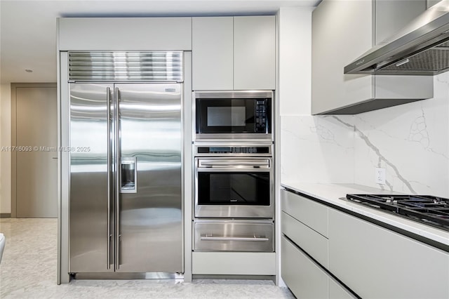 kitchen with light stone countertops, built in appliances, tasteful backsplash, and wall chimney range hood