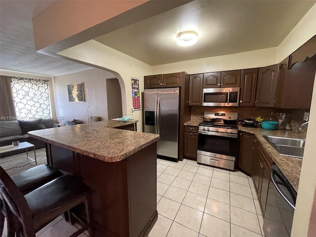 kitchen with stainless steel appliances, light tile patterned flooring, dark brown cabinetry, a sink, and a kitchen bar