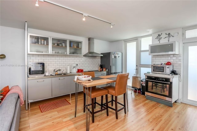 kitchen featuring stainless steel appliances, decorative backsplash, glass insert cabinets, light wood-style floors, and wall chimney exhaust hood