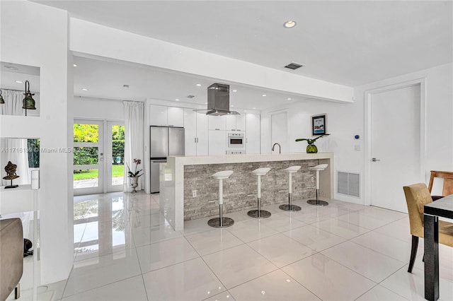 kitchen with stainless steel fridge, french doors, island range hood, light tile patterned floors, and white cabinetry