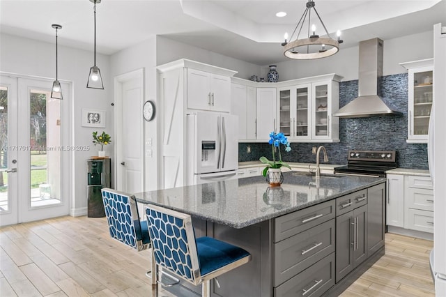 kitchen with white refrigerator with ice dispenser, a center island with sink, wall chimney range hood, sink, and white cabinetry