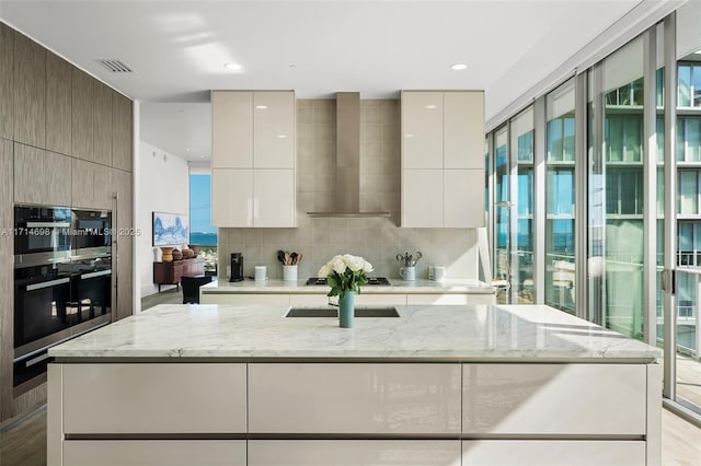 kitchen with wall chimney exhaust hood, light stone counters, double oven, black gas cooktop, and decorative backsplash