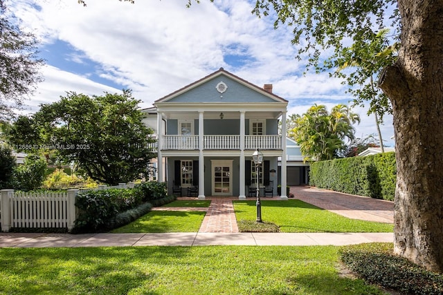 neoclassical / greek revival house with decorative driveway, a chimney, a front yard, fence, and a balcony