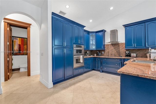 kitchen with blue cabinets, stainless steel double oven, wall chimney range hood, a sink, and visible vents