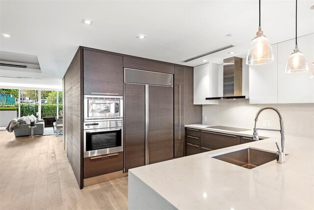 kitchen featuring white cabinetry, sink, wall chimney range hood, built in appliances, and pendant lighting