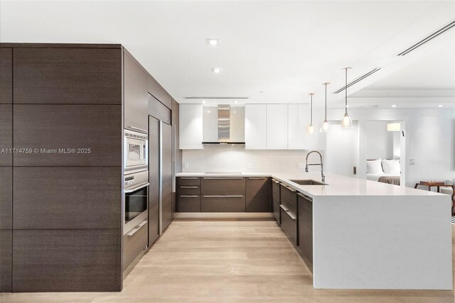 kitchen featuring white cabinets, sink, built in appliances, wall chimney exhaust hood, and decorative light fixtures