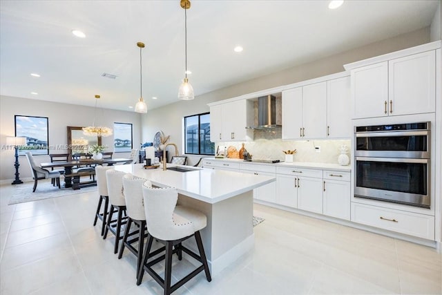 kitchen with wall chimney exhaust hood, double oven, sink, a center island with sink, and white cabinets