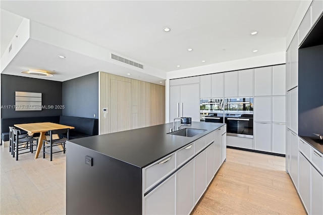 kitchen with white cabinetry, sink, stainless steel double oven, light hardwood / wood-style flooring, and a center island with sink