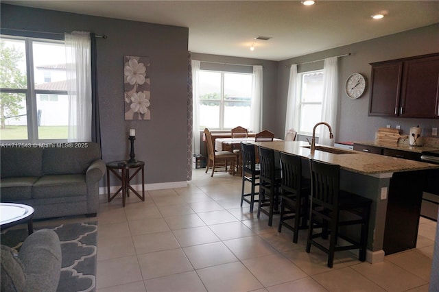 kitchen featuring sink, light stone counters, dark brown cabinets, light tile patterned floors, and a center island with sink