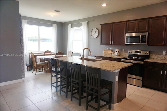 kitchen with a kitchen bar, appliances with stainless steel finishes, light stone counters, sink, and an island with sink