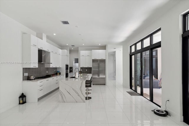 kitchen featuring white cabinets, a center island with sink, wall chimney exhaust hood, tasteful backsplash, and stainless steel built in refrigerator