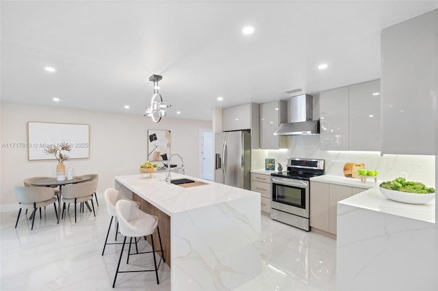 kitchen with a center island with sink, wall chimney range hood, sink, appliances with stainless steel finishes, and decorative light fixtures