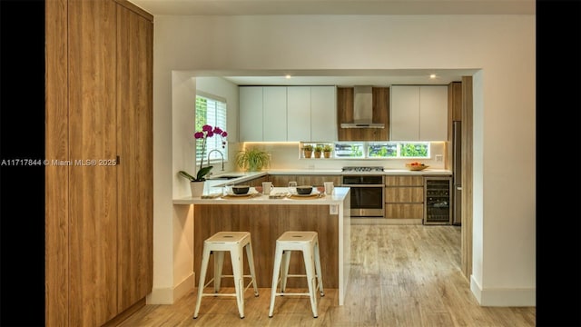 kitchen with kitchen peninsula, wall chimney exhaust hood, stainless steel appliances, beverage cooler, and white cabinetry