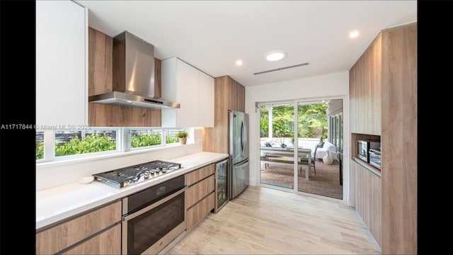 kitchen featuring stainless steel appliances, beverage cooler, wall chimney range hood, light hardwood / wood-style flooring, and white cabinetry