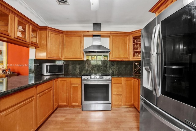 kitchen featuring light wood-type flooring, appliances with stainless steel finishes, dark stone countertops, wall chimney range hood, and crown molding